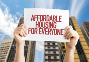 A sign that says "Affordable Housing for Everyone" being held up by two hands with a blue sky and buildings in the background